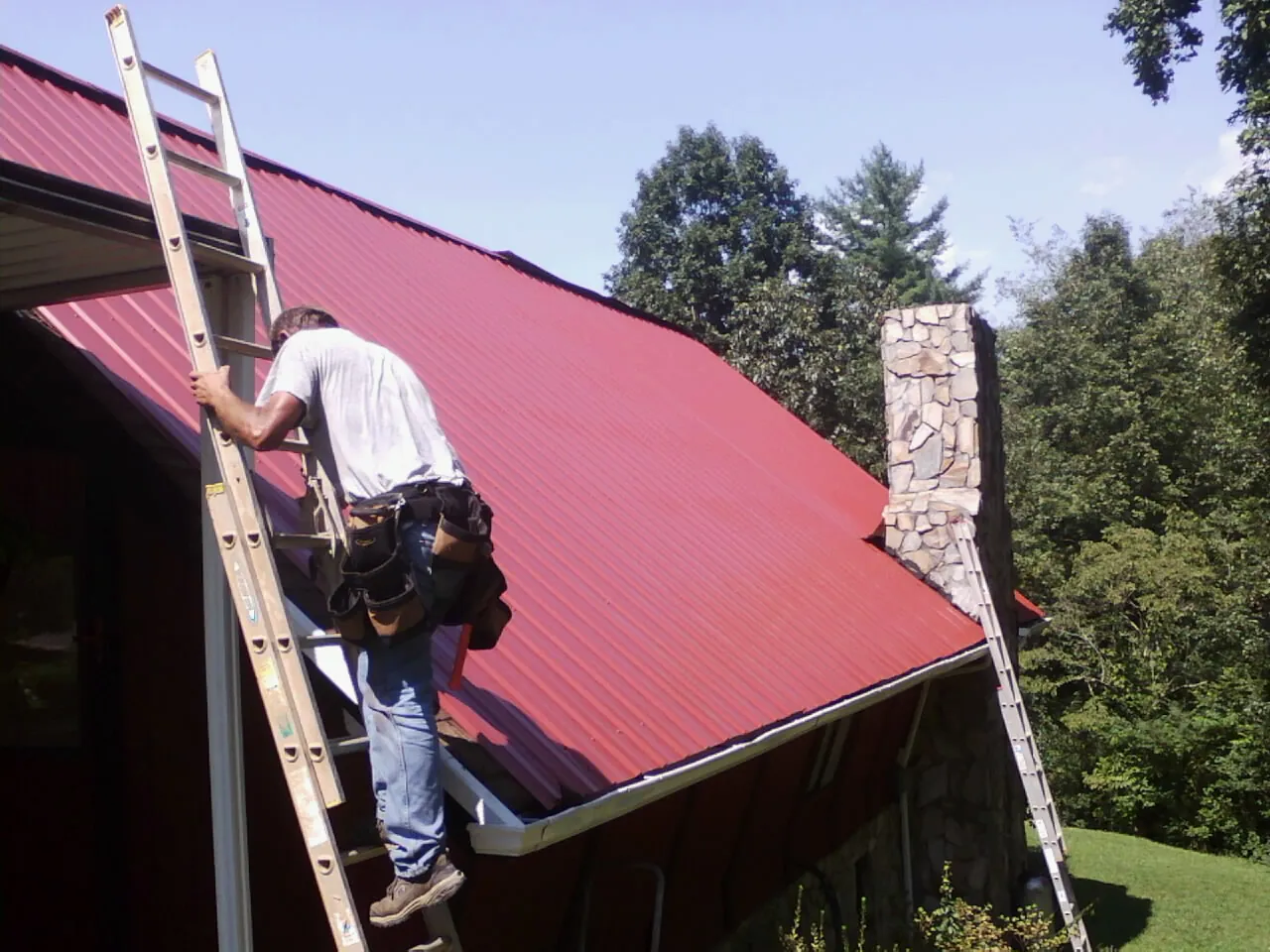 Roofer on a ladder beside a red metal roof and stone chimney — Soffit & Fascia Repair by Roofing Brothers in Doniphan